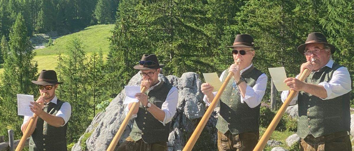 Vier Männer in traditioneller Kleidung spielen lange Holzinstrumente in einer malerischen Außenumgebung. Sie stehen auf einer Grasfläche mit Felsen und Bäumen im Hintergrund. Jeder Mann hält ein Musikblatt in der Hand.