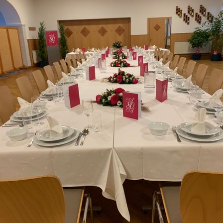 Ein langer Esstisch ist für eine besondere Veranstaltung gedeckt, geschmückt mit roten Blumen, monogrammierten Servietten und Besteck. Der Raum hat Holzwände und -böden, mit einem Heizkörper und Topfpflanzen im Hintergrund.