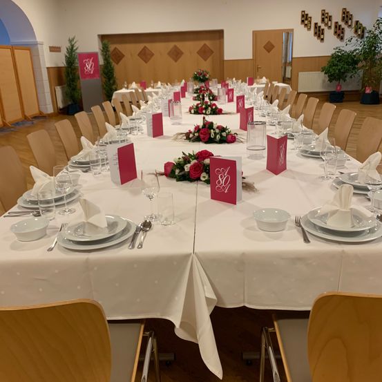 Ein langer Esstisch ist für eine besondere Veranstaltung gedeckt, geschmückt mit roten Blumen, monogrammierten Servietten und Besteck. Der Raum hat Holzwände und -böden, mit einem Heizkörper und Topfpflanzen im Hintergrund.
