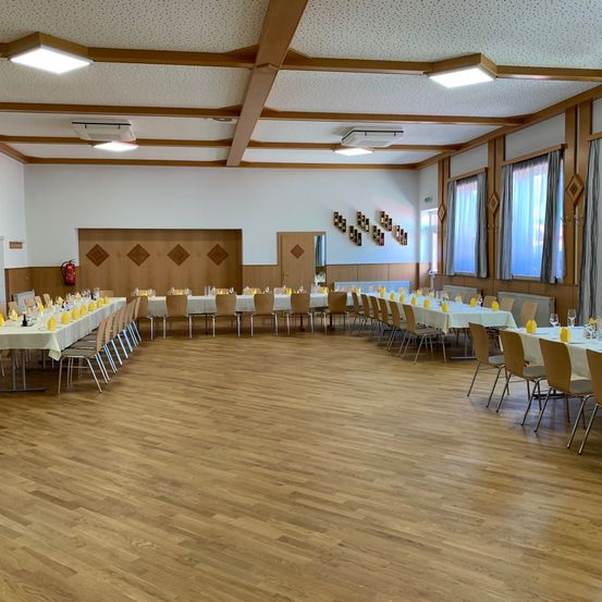 Ein Speisesaal mit Holzboden und weißen Wänden. Der Raum hat mehrere Tische und Stühle, alle in Reihen aufgestellt. Es gibt Lampen an der Decke.