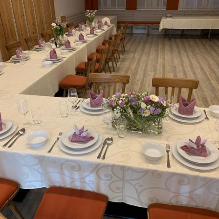 Ein Esszimmer mit mehreren Tischen, jeder mit Tellern, Gläsern, Besteck und Blumen. Die Tische haben gemusterte Tischdecken. Der Raum hat Holzböden und Wände.