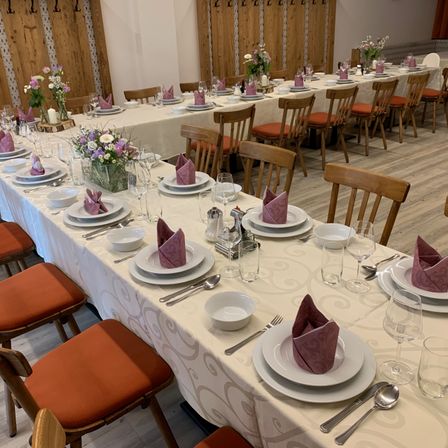 Ein langer Tisch für eine Mahlzeit mit weißen Tellern, lila Servietten, Gläsern und Besteck. Holzstühle mit orangefarbenen Kissen sind um den Tisch herum angeordnet. Holzwände und Blumen in Vasen sind im Hintergrund.