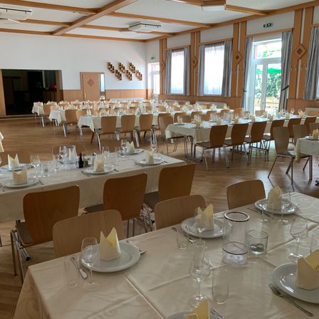 Ein Innenspeisesaal ist mit zahlreichen Tischen eingerichtet, die jeweils mit Tellern, Weingläsern und Servietten ordentlich arrangiert sind. Der Saal ist geräumig mit Holzböden und großen Fenstern, die für eine helle Atmosphäre sorgen.
