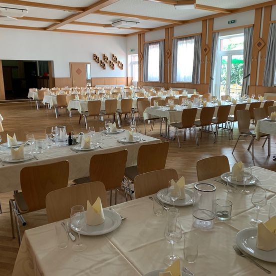 Ein Innenspeisesaal ist mit zahlreichen Tischen eingerichtet, die jeweils mit Tellern, Weingläsern und Servietten ordentlich arrangiert sind. Der Saal ist geräumig mit Holzböden und großen Fenstern, die für eine helle Atmosphäre sorgen.
