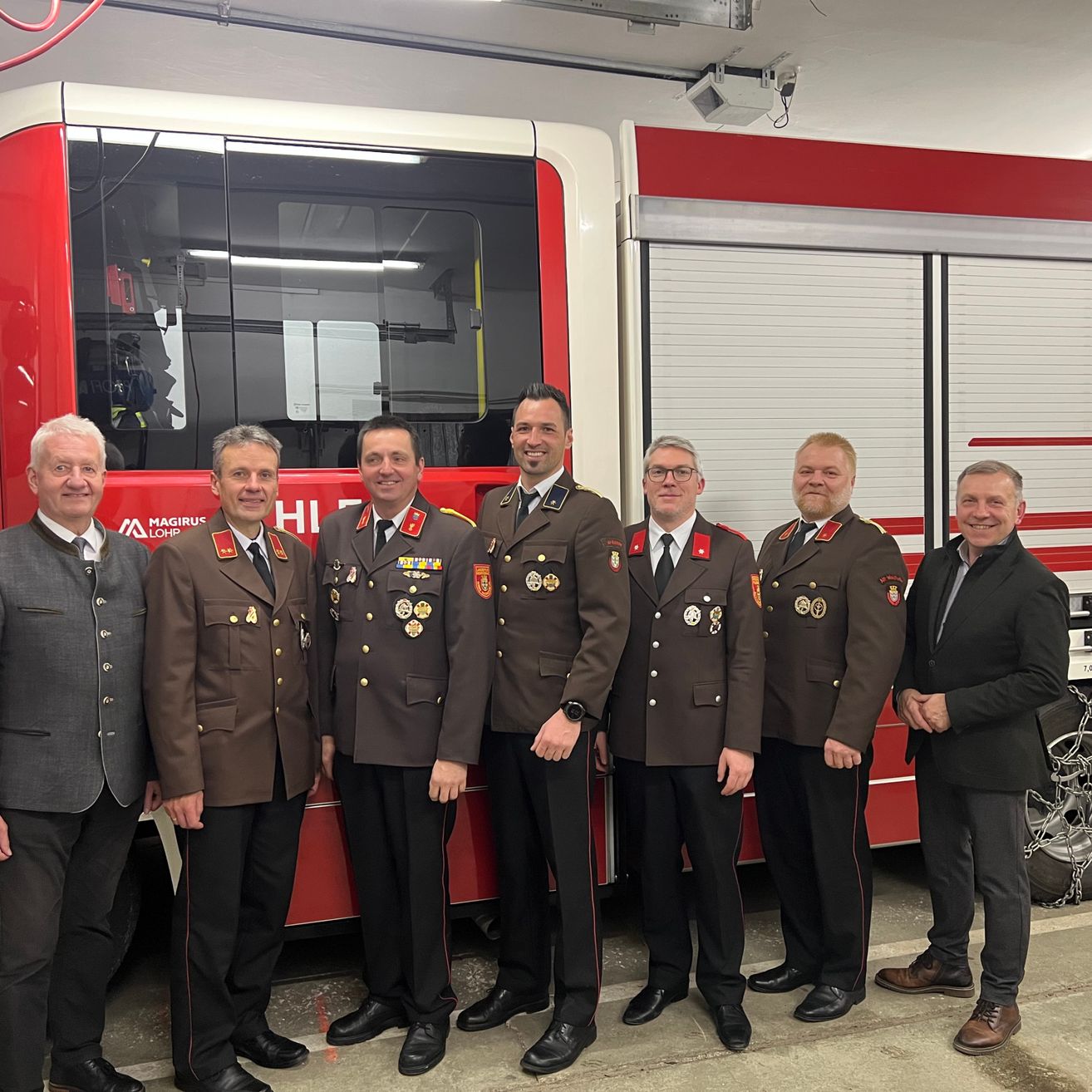 Sieben Männer in Uniformen stehen neben einem Feuerwehrauto. Sie alle lächeln und schauen in die Kamera. Ein Mann trägt eine Uhr.