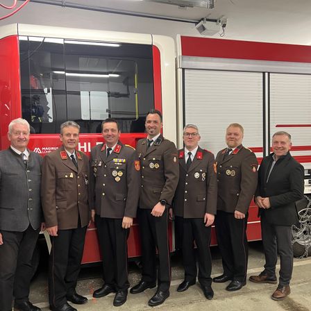 Sieben Männer in Uniformen stehen neben einem Feuerwehrauto. Sie alle lächeln und schauen in die Kamera. Ein Mann trägt eine Uhr.