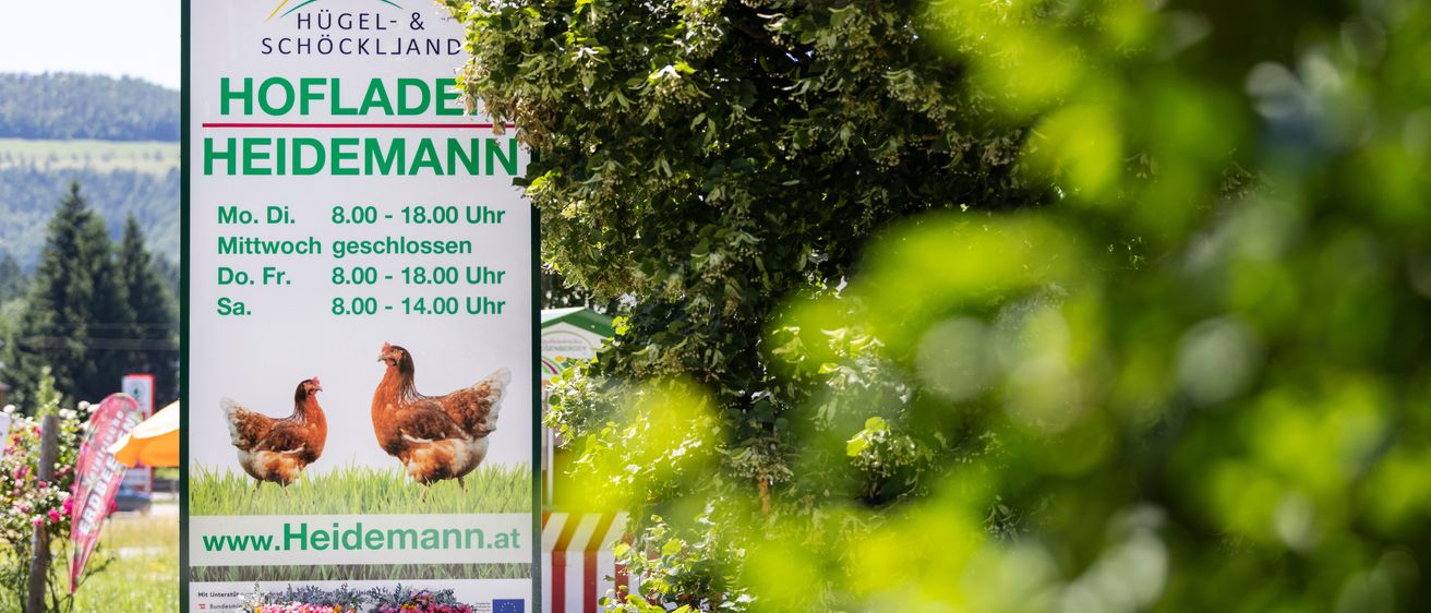 Ein Schild wirbt für einen Bauernhof mit zwei Hühnern und einer Website-Adresse. Es listet Öffnungszeiten für verschiedene Tage auf.