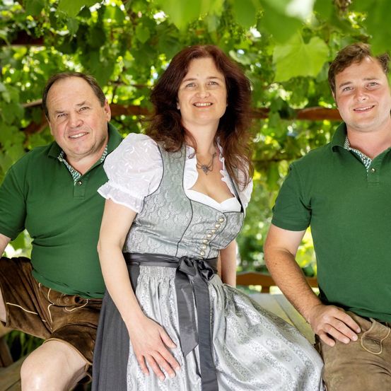 Drei Personen posieren für ein Foto in einem Garten mit grünen Blättern. Eine Frau in traditioneller Kleidung steht zwischen zwei Männern, alle lächeln.