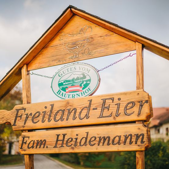 Ein Holzschild für Freiland-Eier Farm Heidmann. Ein grünes rundes Schild mit Logo und Text steht für "Gutes vom Bauernhof". Das Schild ist an einer Holzstruktur montiert.