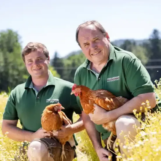 Zwei Männer in grünen Poloshirts sitzen in einem Feld, jeder hält ein Huhn. Der Mann auf der linken Seite lächelt und hält ein Huhn auf dem Schoß. Der Mann auf der rechten Seite hält ein Huhn in den Armen und lächelt.