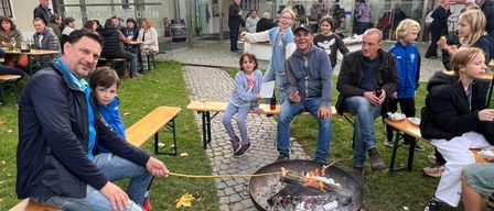 A group of people are sitting around a fire pit, some roasting marshmallows. A man holds a stick with a marshmallow. Nearby, a woman with a child smiles. Behind them, a building with a glass roof stands.