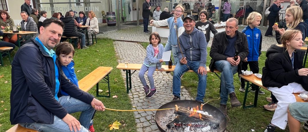 Eine Gruppe von Menschen sitzt um ein Feuer herum, einige rösten Marshmallows. Ein Mann hält einen Stock mit einem Marshmallow. In der Nähe lächelt eine Frau mit einem Kind. Dahinter steht ein Gebäude mit einem Glasdach.