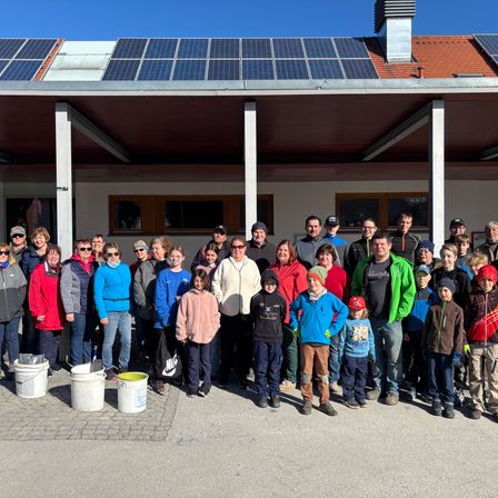 Eine Gruppe von Menschen steht vor einem Gebäude mit Solarmodulen, lächelt und posiert für ein Foto. Eimer stehen vor ihnen auf dem Boden.