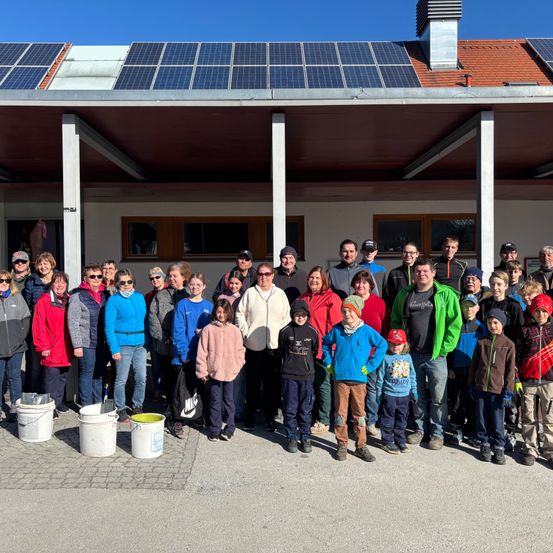 A group of people stand outside a building with solar panels, smiling and posing for a photo. Buckets are placed on the ground in front of them.