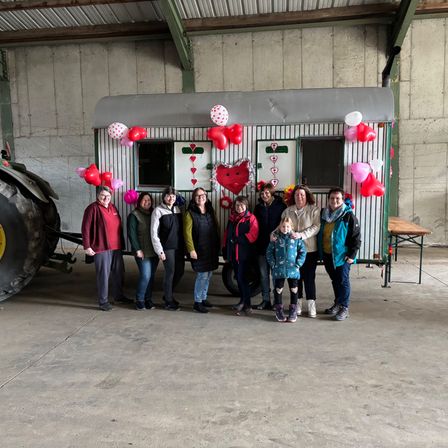 Eine Gruppe von Menschen, einschließlich Kindern, posiert vor einem dekorierten Wohnwagen mit Luftballons und Herzmotiven. Sie befinden sich in einer Betongarage.