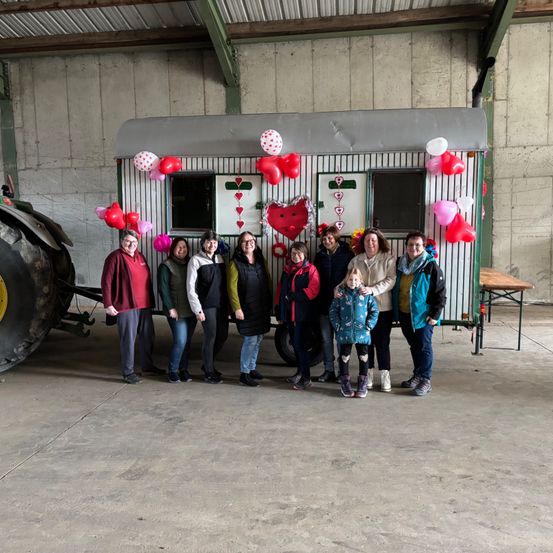 A group of people, including children, poses in front of a decorated caravan with balloons and heart motifs. They are in a concrete garage.