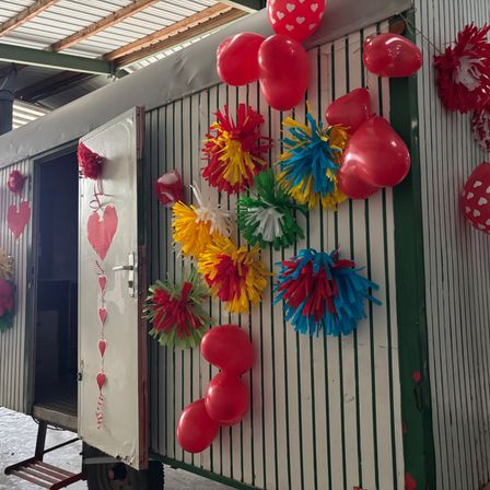 Eine weiße und grüne Struktur mit roten Luftballons und bunten Wimpelgirlanden, mit einer offenen Tür auf der linken Seite. Die Luftballons sind in verschiedenen Farben, einschließlich rot, gelb und blau. Die Wimpelgirlanden sind in verschiedenen Farben, einschließlich rot, blau und gelb. Die Struktur hat ein grünes und weißes Streifendesign.