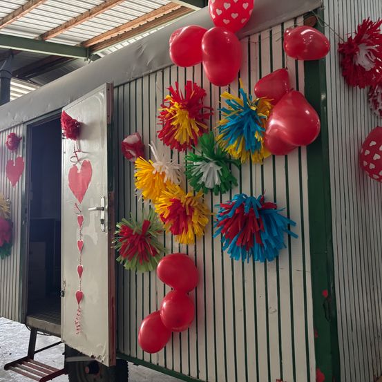 A white and green structure with red balloons and colorful pom-poms, with an open door on the left side. The balloons are in various colors, including red, yellow, and blue. The pom-poms are in different colors, including red, blue, and yellow. The structure has a green and white striped design.