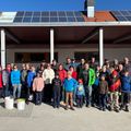 Eine große Gruppe von Menschen in Winterkleidung steht vor einem Gebäude mit Solarmodulen auf dem Dach. Sie lächeln und scheinen für ein Foto zu posieren.
