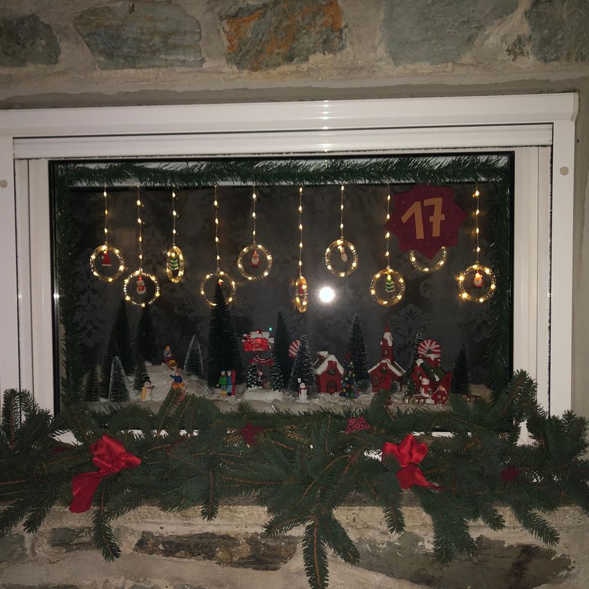 Ein Fensterdisplay mit Weihnachtslichtern, Ornamenten und einer festlichen Szene. Der Fensterrahmen ist weiß, und die Dekorationen umfassen Kiefernzweige mit roten Bändern. Es gibt Miniatur-Weihnachtsbäume und Häuser, und die Nummer 17 ist prominent angebracht.