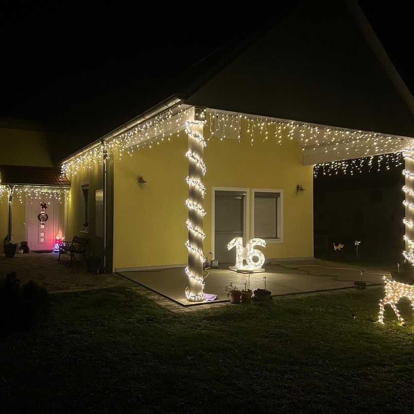 Ein Haus ist nachts mit Weihnachtslichtern beleuchtet, darunter eine Nummer 16 und eine Rentierdekoration. Das Haus hat eine Veranda mit einer Bank und Topfpflanzen.