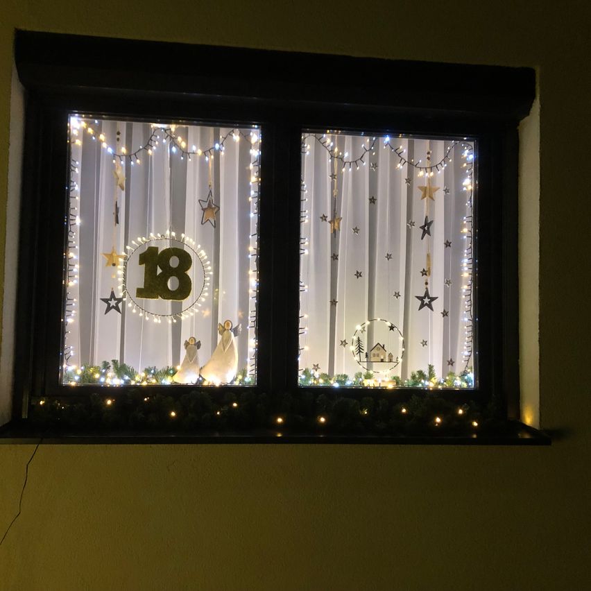 Ein Fenster, das für eine Geburtstagsfeier dekoriert ist. Das Fenster hat zwei Abschnitte mit weißen Vorhängen, die jeweils mit goldenen Sternen und Weihnachtslichtdekorationen geschmückt sind. Der linke Abschnitt zeigt eine große goldene Zahl 18 mit zwei Engelsfiguren. Der rechte Abschnitt zeigt ein Haus und eine kreisförmige Dekoration.