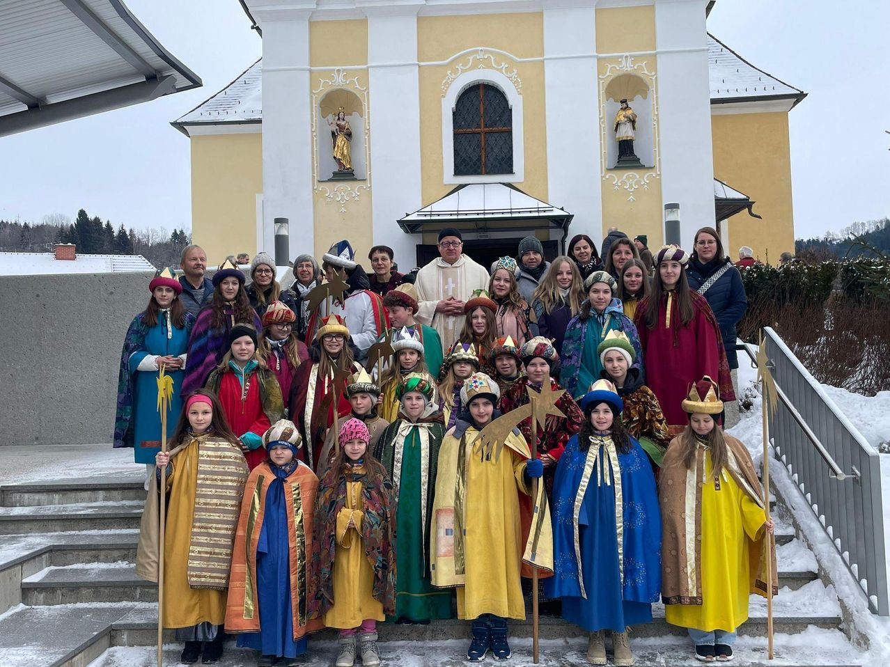Eine Gruppe von Kindern und Erwachsenen in farbenfrohen Kostümen posiert für ein Foto vor einer Kirche mit Statuen an den Wänden.