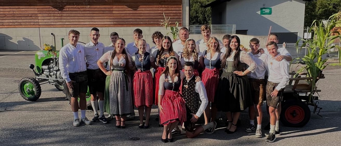 Eine Gruppe von Menschen in traditioneller bayerischer Kleidung, die für ein Foto vor einem Gebäude mit Bergen im Hintergrund posiert.