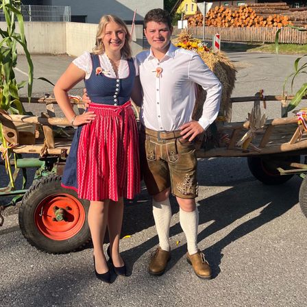 Ein Paar steht neben einem mit Mais und Heu beladenen Holzwagen, gekleidet in traditionelle bayerische Kleidung, mit einem Haus und einem Holzstapel im Hintergrund.