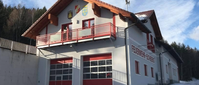 Ein weißes Gebäude mit braunem Dach und roten Akzenten, beschriftet mit 'Feuerwehr-Riedenberg', steht auf einer verschneiten Straße mit Balkon und großen Garagentoren.