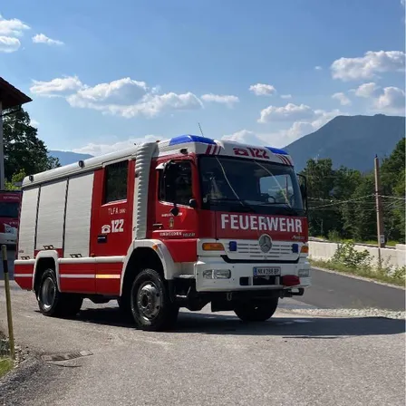 Ein rot-weißer Feuerwehrwagen mit 'FEUERWEHR' auf der Front fährt auf einer Straße. Der Himmel ist blau mit Wolken, und im Hintergrund sind Berge zu sehen.