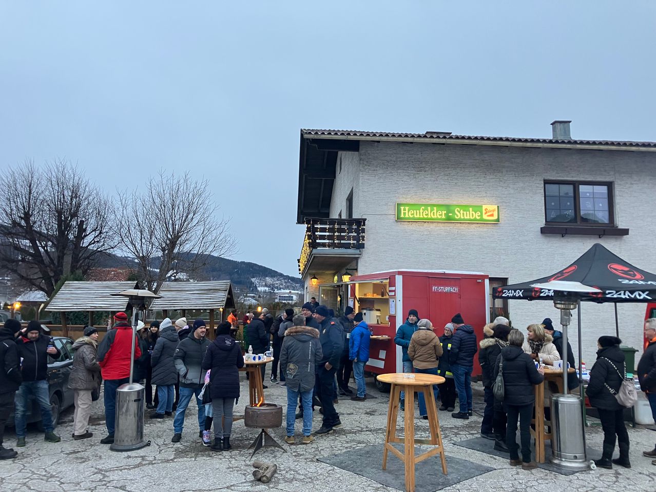 Eine Gruppe von Menschen steht um Tische herum vor einem Gebäude namens Heufelder-Stube. Es gibt Heizkörper und ein Essensmobil. Der Himmel ist bewölkt und Berge sind in der Ferne.