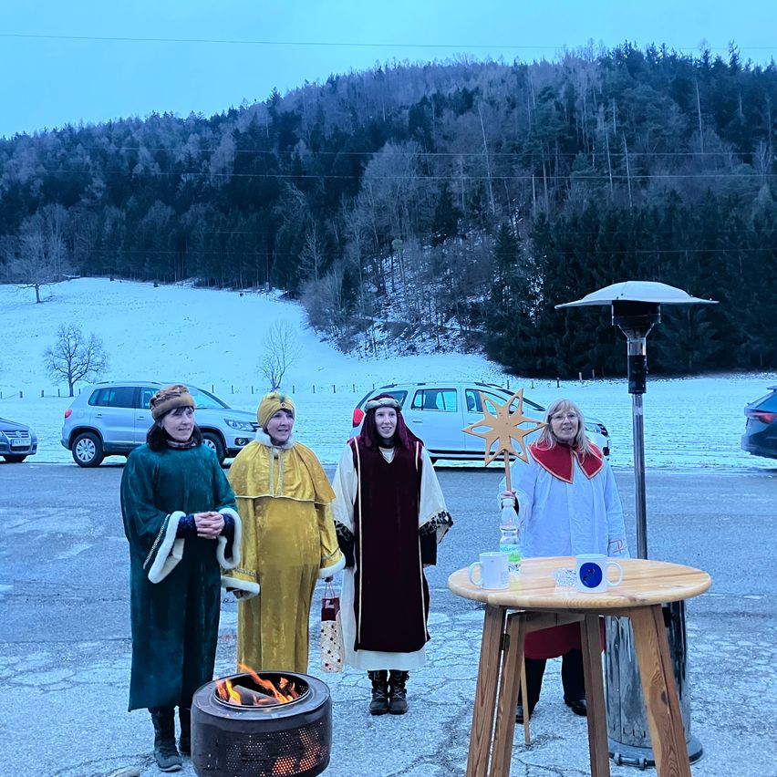 Vier Personen in traditioneller Kleidung stehen neben einem Feuerkorb auf einem verschneiten Parkplatz. Ein Holztisch hält Tassen und Flaschen, mit einem Stern und einem Auto im Hintergrund.
