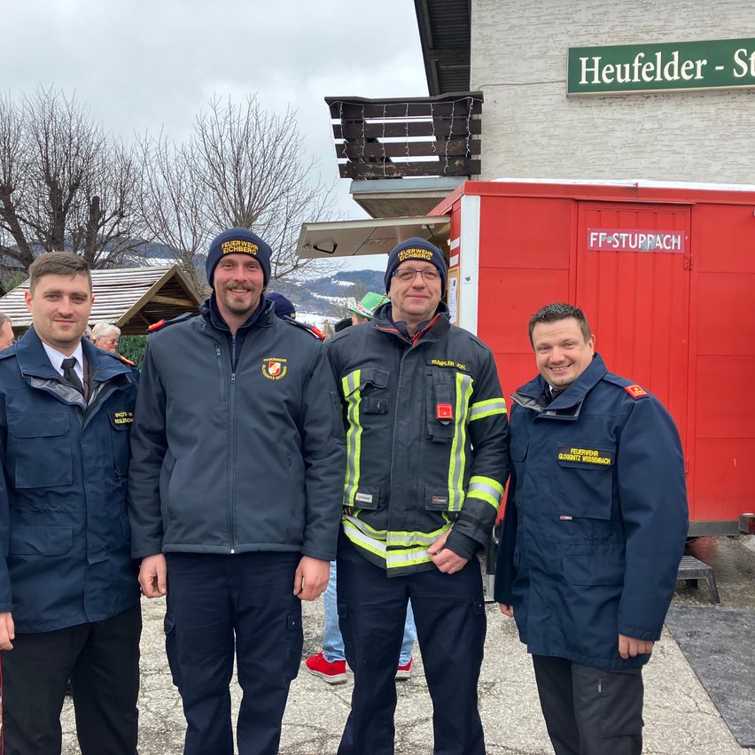 Vier Männer stehen vor einem Gebäude mit einem Schild, das Heufelder-St. trägt. Die beiden in der Mitte tragen Feuerwehruniformen. Sie scheinen für ein Foto zu posieren.