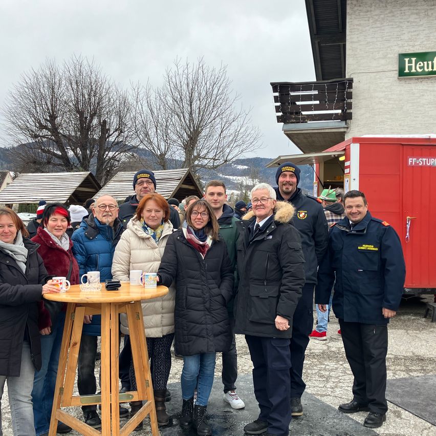 Eine Gruppe von Menschen steht vor einem Gebäude mit einem roten Container, der mit Heuf beschriftet ist. Sie tragen Winterkleidung und einige haben Mützen. Ein Tisch mit Tassen steht in der Mitte. Hinter ihnen sind Bäume und ein Berg.