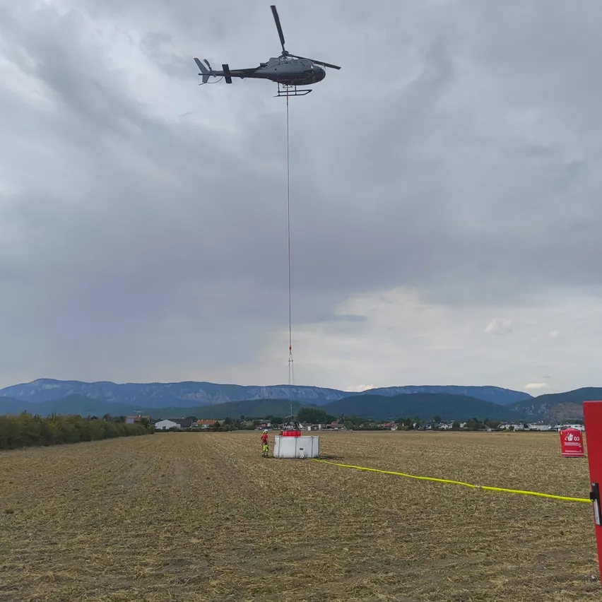 Ein Hubschrauber schwebt über einem Feld und senkt einen Container mit einem Seil. Eine Person in einer gelben Weste steht in der Nähe. Berge sind in der Ferne sichtbar.