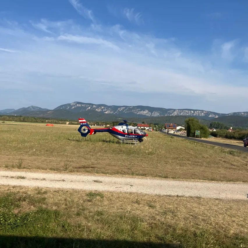 Ein Polizeihubschrauber ist auf einem Grasfeld mit Bergen im Hintergrund unter einem klaren blauen Himmel geparkt.