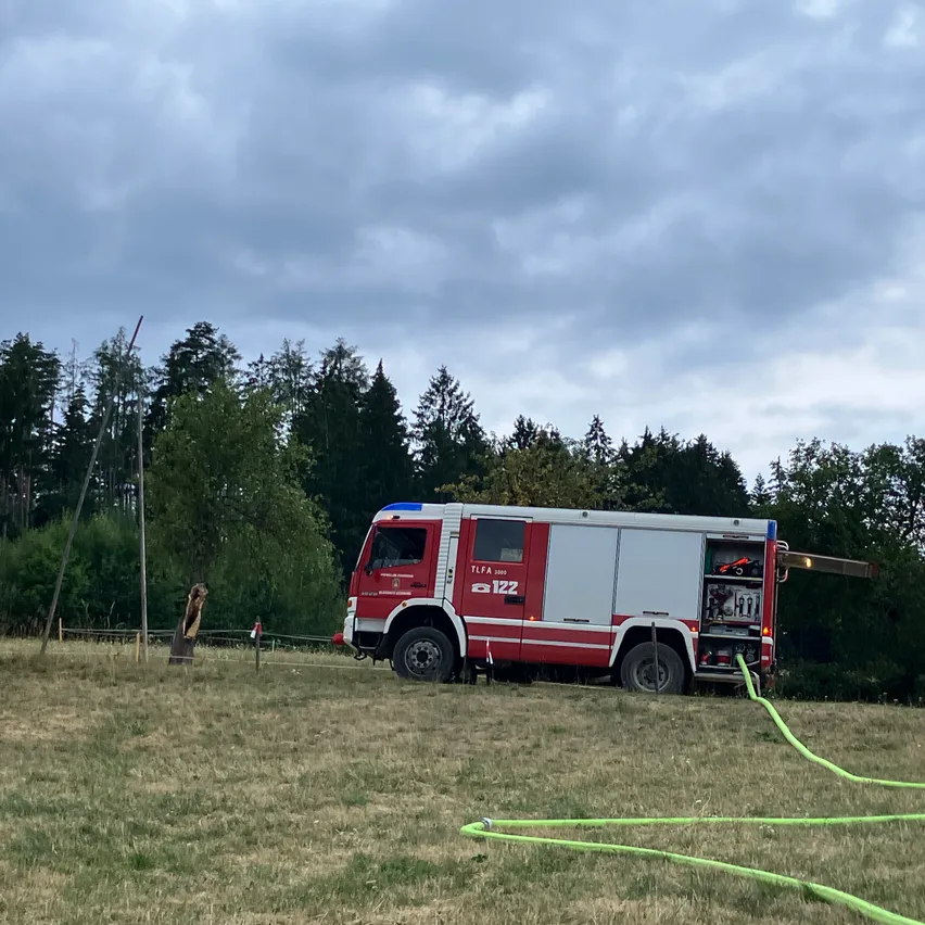 Ein rot-weißer Feuerwehrwagen steht auf einer Wiese mit Bäumen im Hintergrund. Am Wagen ist ein grüner Schlauch angeschlossen.