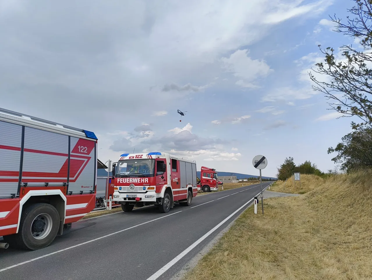 Ein roter Feuerwehrwagen mit der Nummer 122 fährt auf der Straße, mit einem weiteren Feuerwehrwagen und einem Hubschrauber am Himmel.