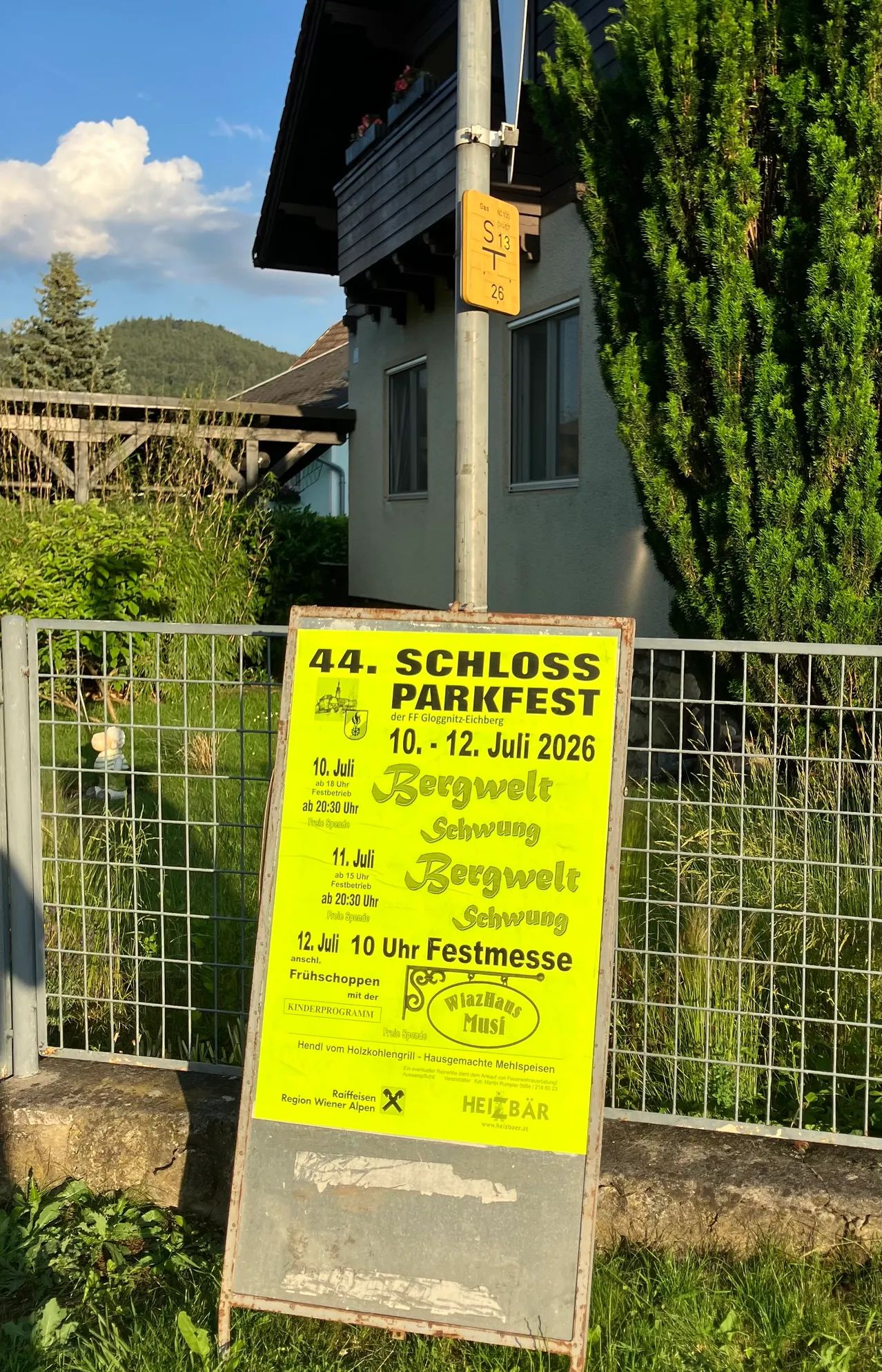 Ein gelbes Schild an einem Zaun bewirbt das 44. Schloss Parkfest vom 10. bis 12. Juli 2026. Die Veranstaltung zeigt einen Berghintergrund, ein Gebäude und üppiges Grün.