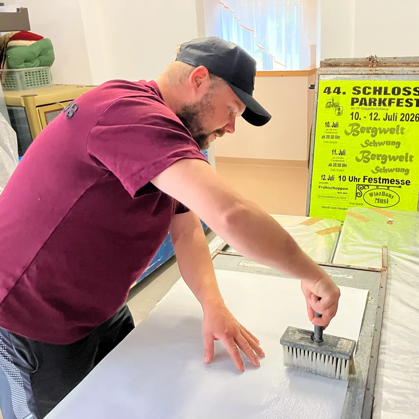 Ein Mann in einem purpurroten T-Shirt und einer schwarzen Kappe arbeitet mit einem Pinsel auf einer weißen Oberfläche. Er bereitet wahrscheinlich etwas vor. Dahinter befindet sich ein gelbes Plakat.
