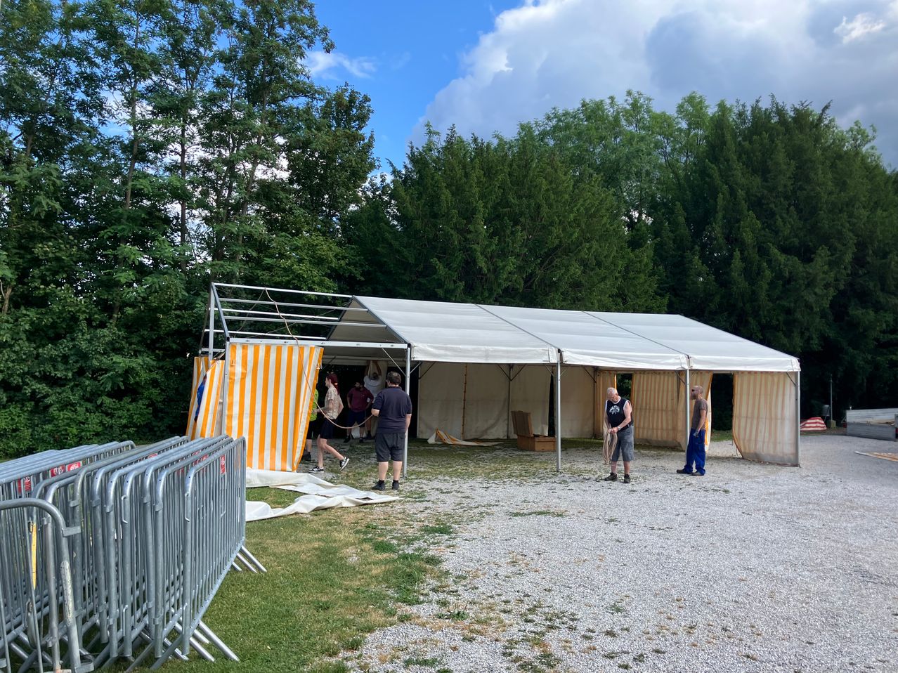 People set up a large tent in a field with gravel ground. A group of individuals work on the tent's entrance.