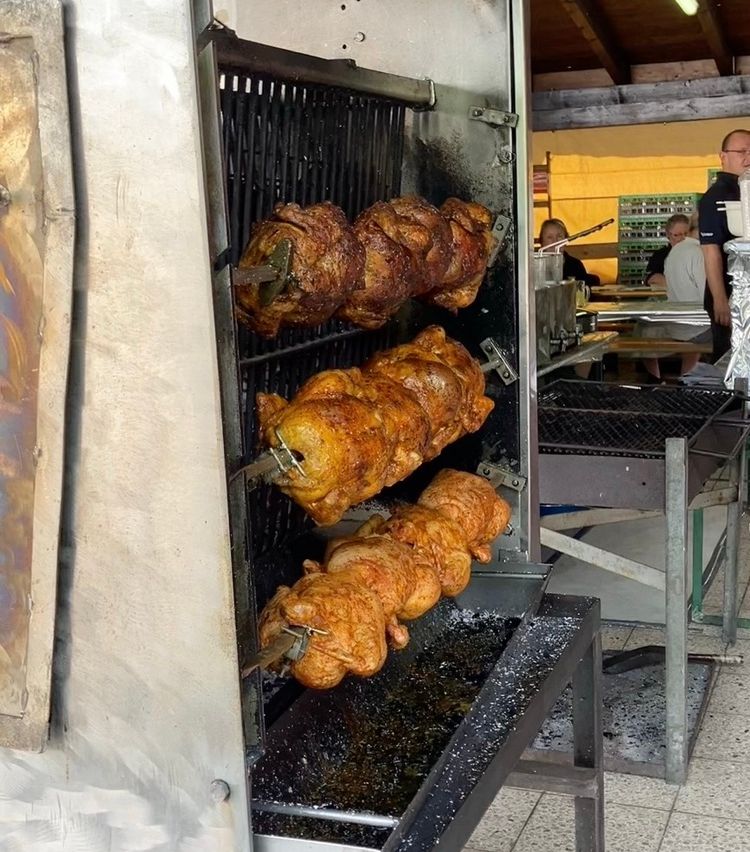 A close-up view of a rotisserie oven with three chickens being cooked. The oven is metal with a glass door. In the background, people are standing and sitting.