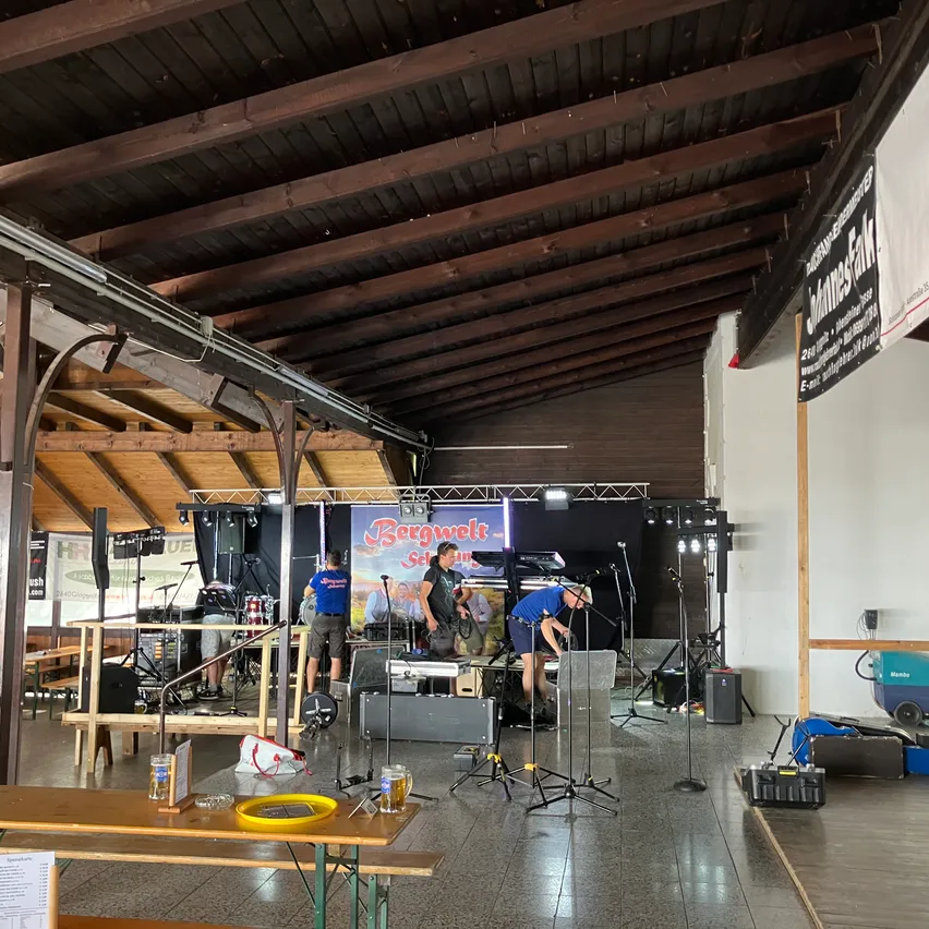 Eine Band richtet auf einer Bühne mit Musikinstrumenten und Ausrüstung in einer großen Halle ein. Holzbalken stützen die Decke, und ein Banner hängt an der Wand.