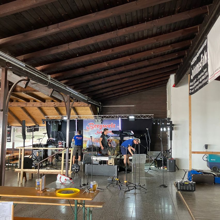 A band is setting up on a stage with musical instruments and equipment in a large indoor venue. Wooden beams support the ceiling, and a banner is hanging on the wall.