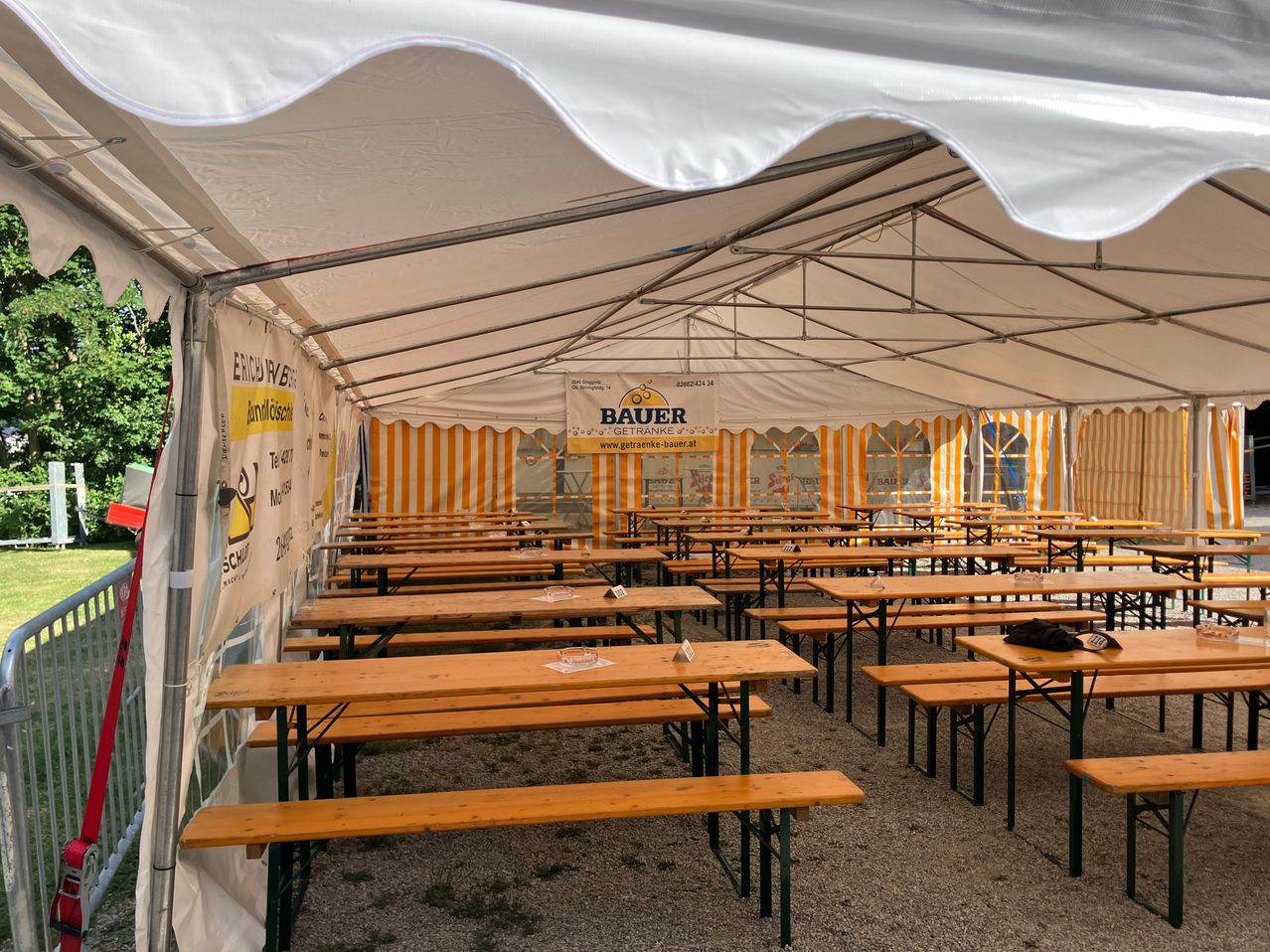 A large tent with a white canopy and yellow and white striped walls. Inside, there are rows of wooden tables and benches, some with glasses and papers on them. A banner with the word Bauer is hanging in the middle.