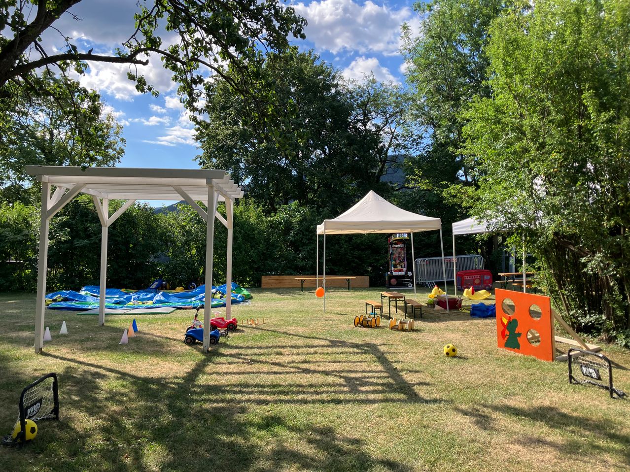 A playground setup with a white gazebo, a white tent, and various toys and games on a grassy field under a partly cloudy sky.
