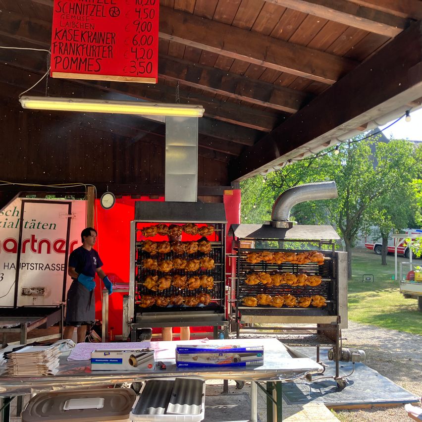 A man stands behind a food stall with two grills cooking chicken. A red sign with prices is above.