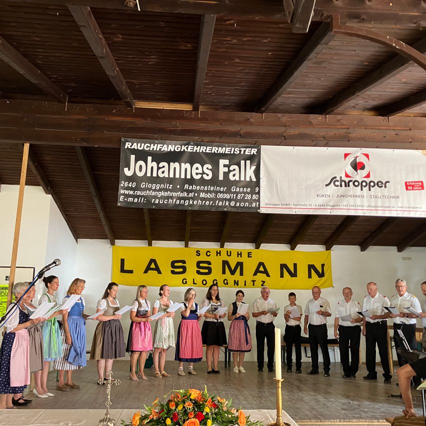 A group of people dressed in traditional attire stand on a stage with a banner reading 'Schuhe Lassmann Gloggnitz'. They hold papers and candles, and a microphone is on the left. Behind them, banners for 'Johannes Faik' and 'Schropper' are visible.