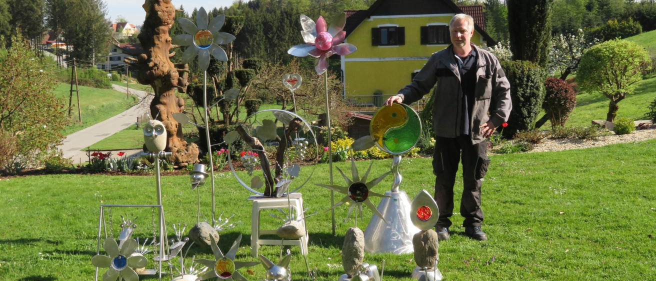Ein Mann steht in einem Garten voller einzigartiger Skulpturen. Hinter ihm ist ein gelbes Haus und üppige Bäume sichtbar. Die Skulpturen umfassen einen Baum mit Hut, Blumen und einen runden Gegenstand.